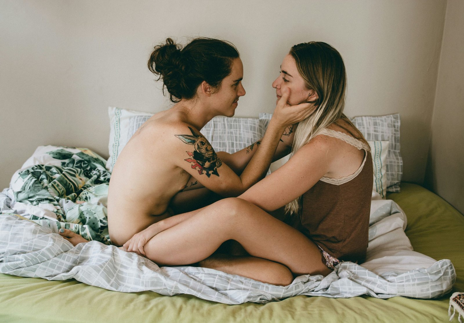 Two people with light skin sit closely together on a bed, gazing into each other’s eyes with tenderness. One person, with tattoos and hair in a loose bun, gently holds the other’s face, who has long blonde hair and is wearing a soft brown tank top. The intimate moment is set against a cozy backdrop of green and white bedding, evoking warmth, connection, and affection.