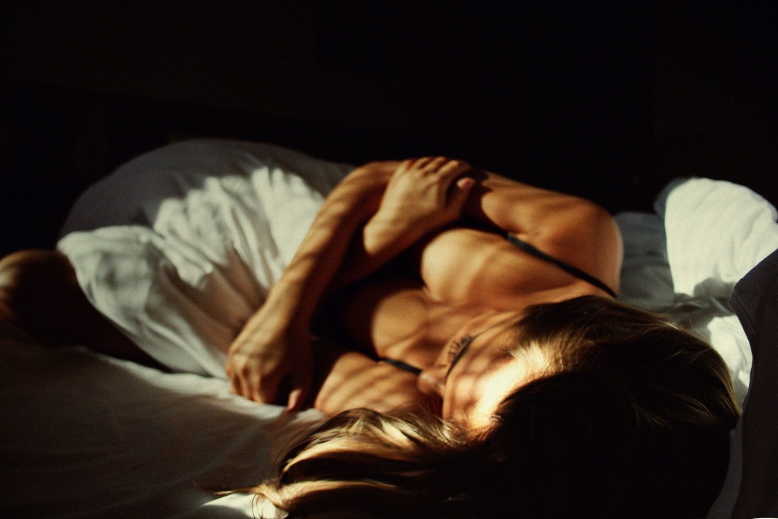 A woman with sun-kissed skin lies on a bed, partially covered by white sheets, bathed in warm, golden light. Shadows from a nearby window create soft patterns across her body. Her posture is relaxed, with one arm gently resting across her chest, evoking a sense of calm, intimacy, and restfulness.